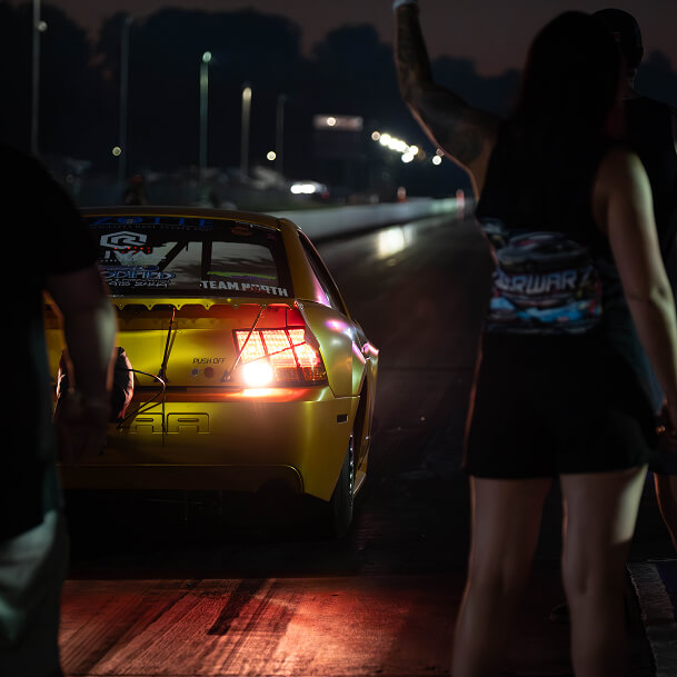 Yellow racing car at night on drag strip with woman raising arm and man standing nearby with Streetcar Addiction