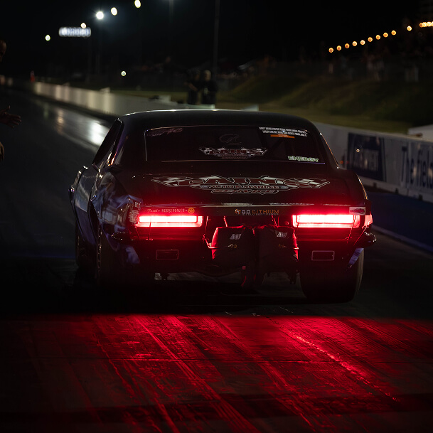 Rear view of a dark-colored muscle car with glowing red brake lights on a drag strip at night with Streetcar Addiction