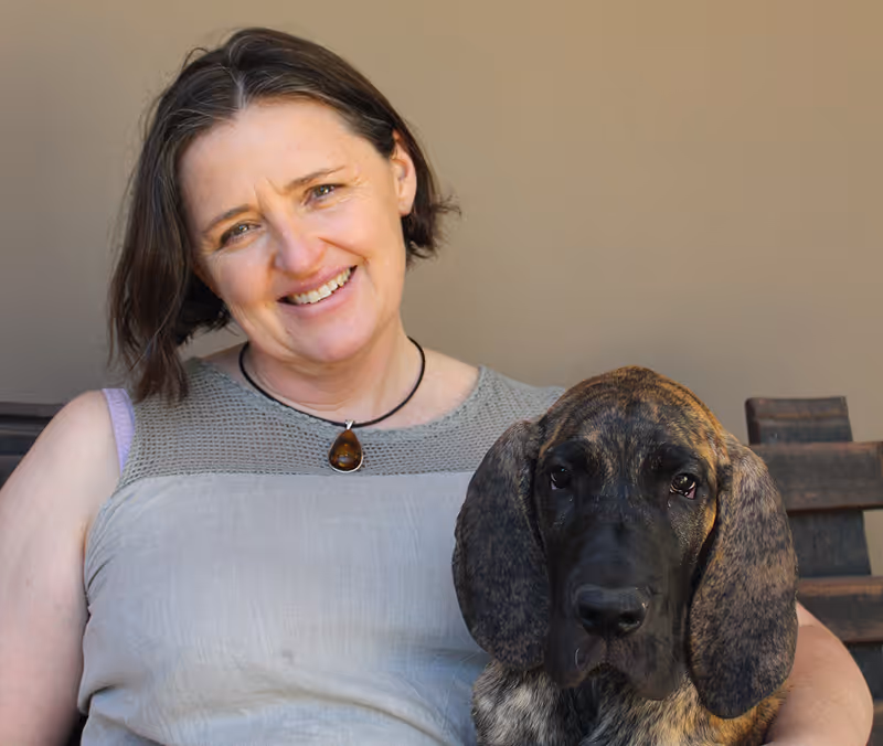 Smiling woman with short brown hair sitting next to a large brindle dog with long ears at citivet bothasig, waiting to welcome new bookings