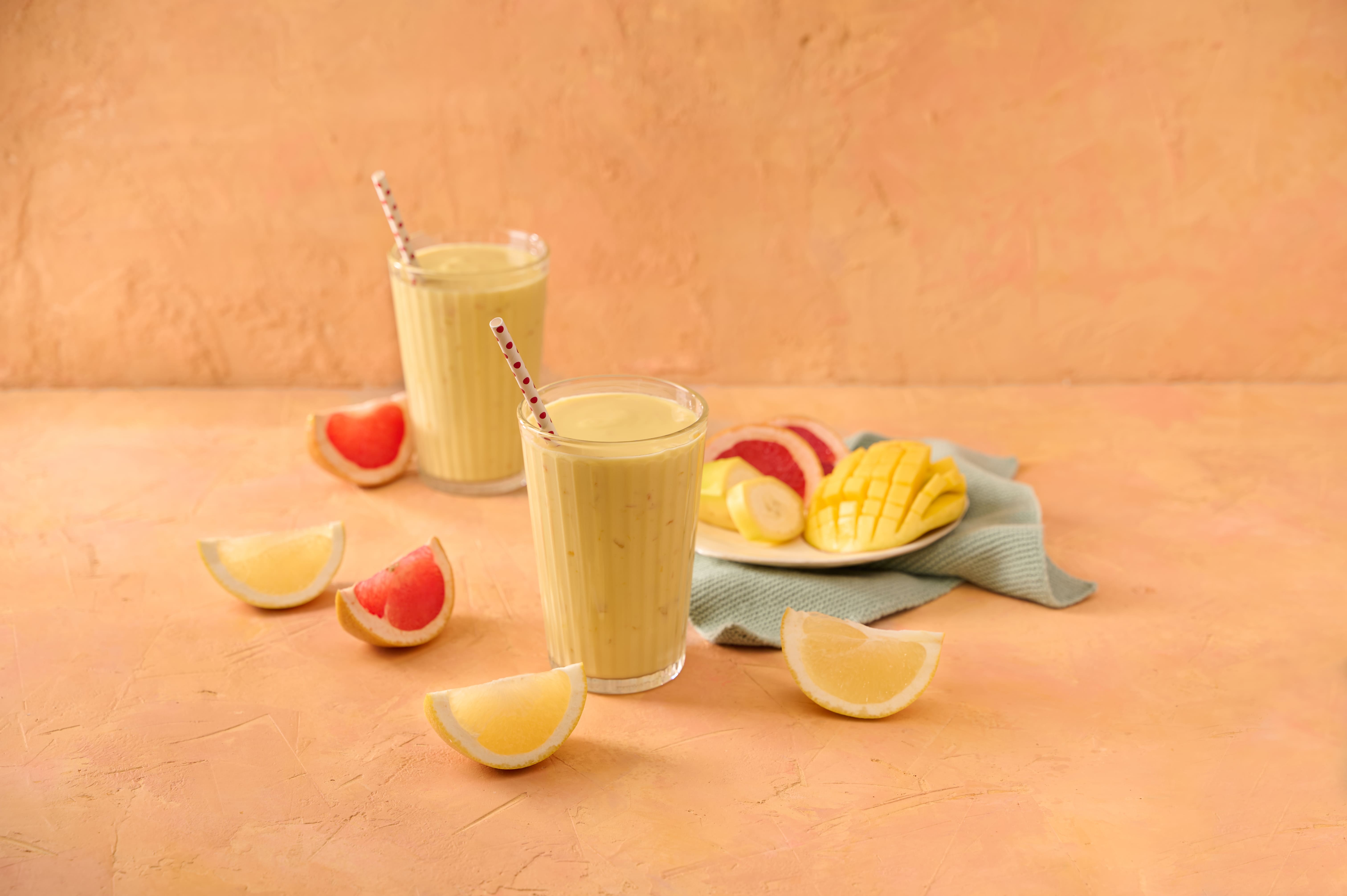 Two glasses of mango smoothie with straws next to fresh mango pieces and grapefruit wedges