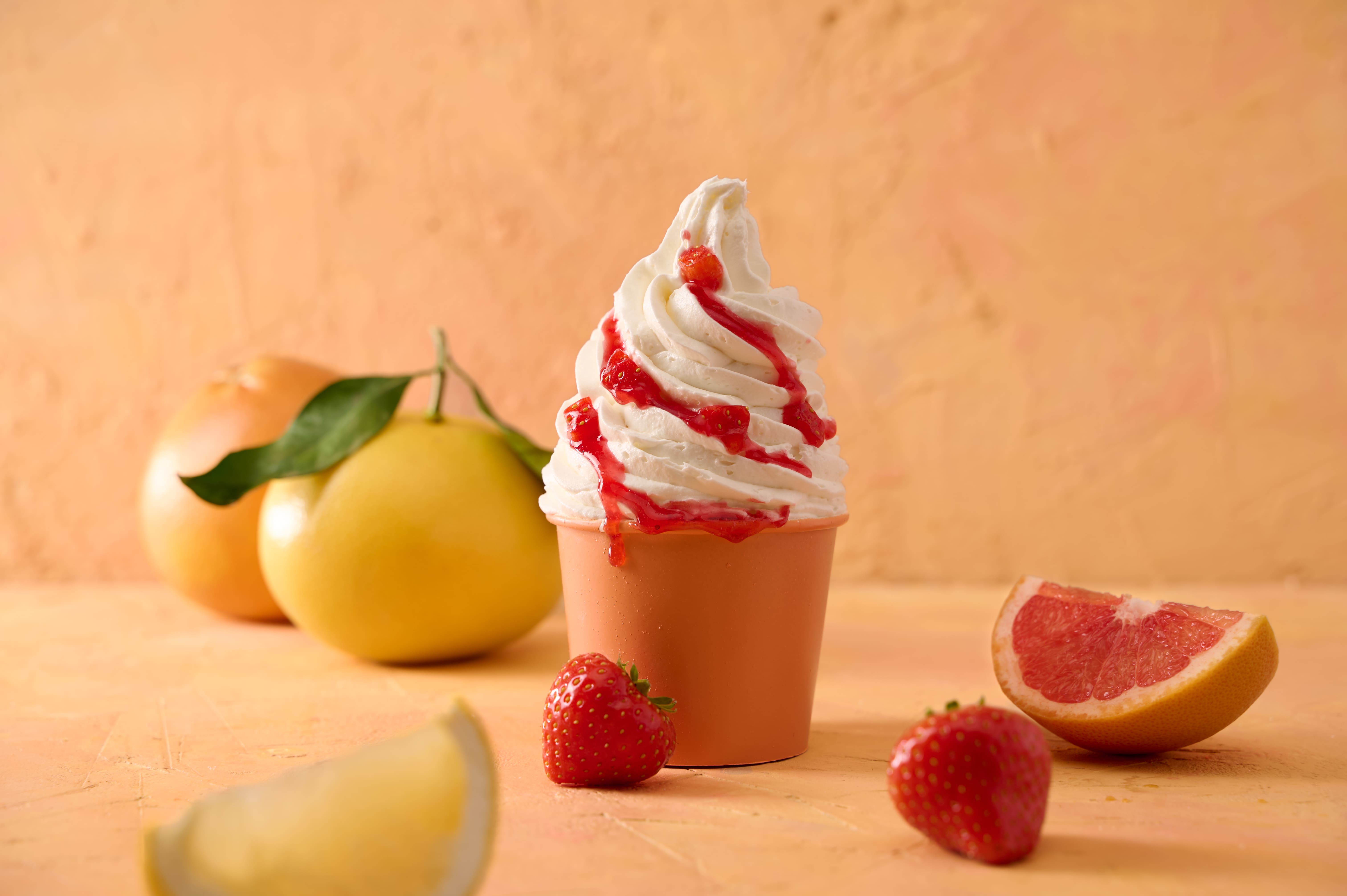 Soft-serve ice cream in a cup drizzled with red sauce, next to grapefruit and strawberries