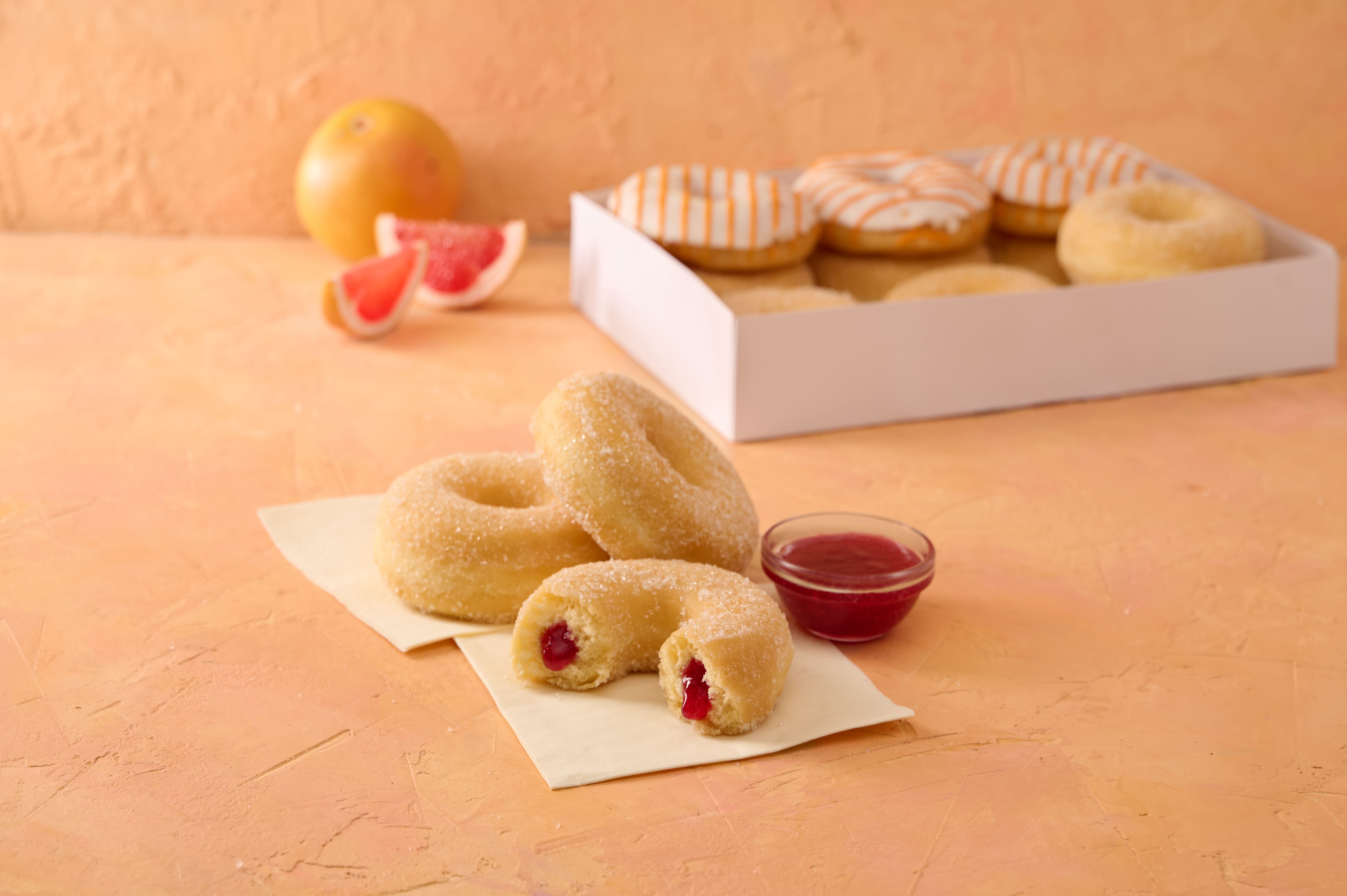 Sugared jam doughnuts on paper next to a bowl of raspberry sauce, a box of glazed doughnuts and grapefruit in the background