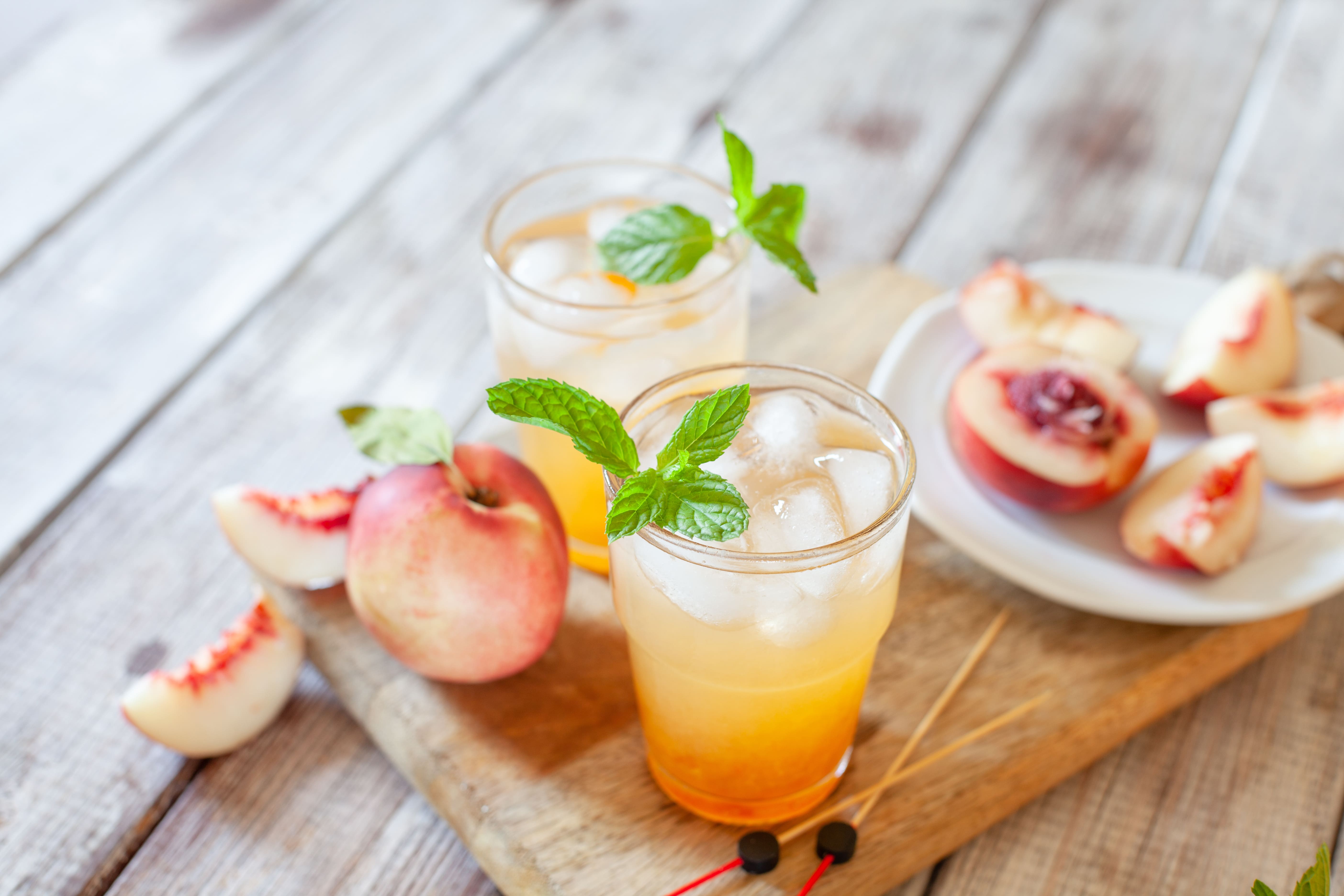 Two glasses of iced peach tea with mint on a wooden board next to fresh peach slices
