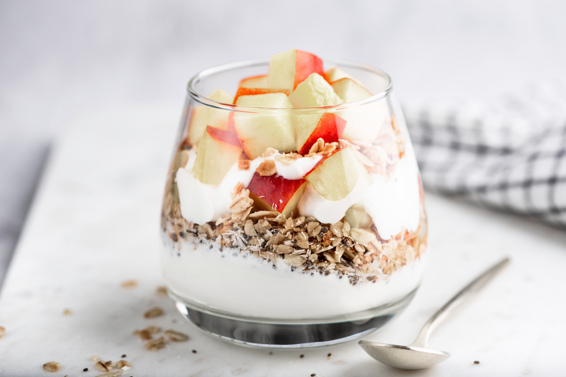 Glass of yogurt parfait with granola and white peach pieces on top, spoon beside it