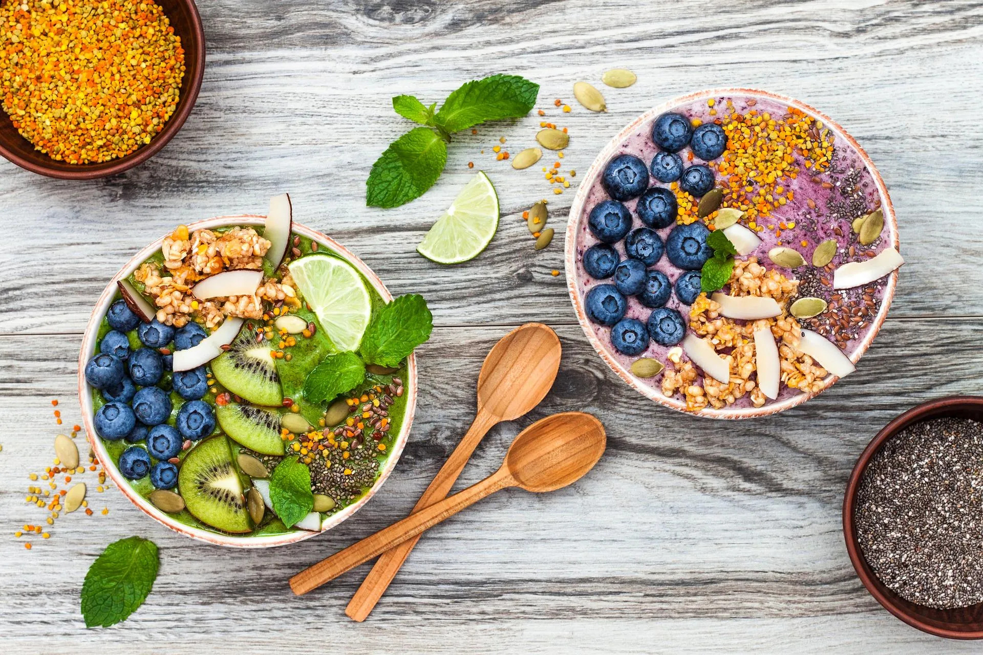 Zwei Smoothie-Bowls mit Beeren, Kiwi, Granola und Samen, von oben mit Holzlöffeln fotografiert