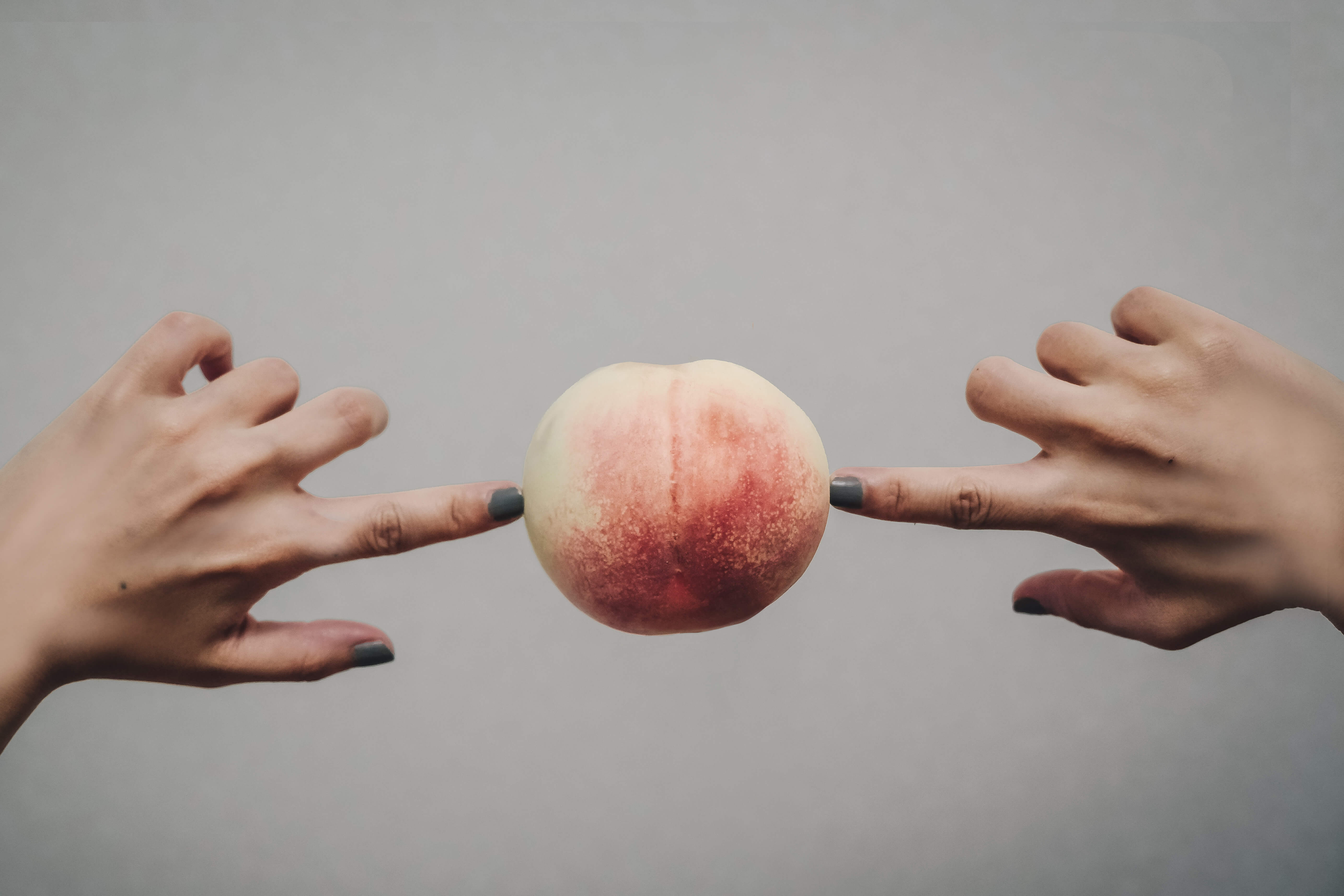 Weiße Pfirsichfrucht zwischen zwei Fingern vor neutralem Hintergrund.“