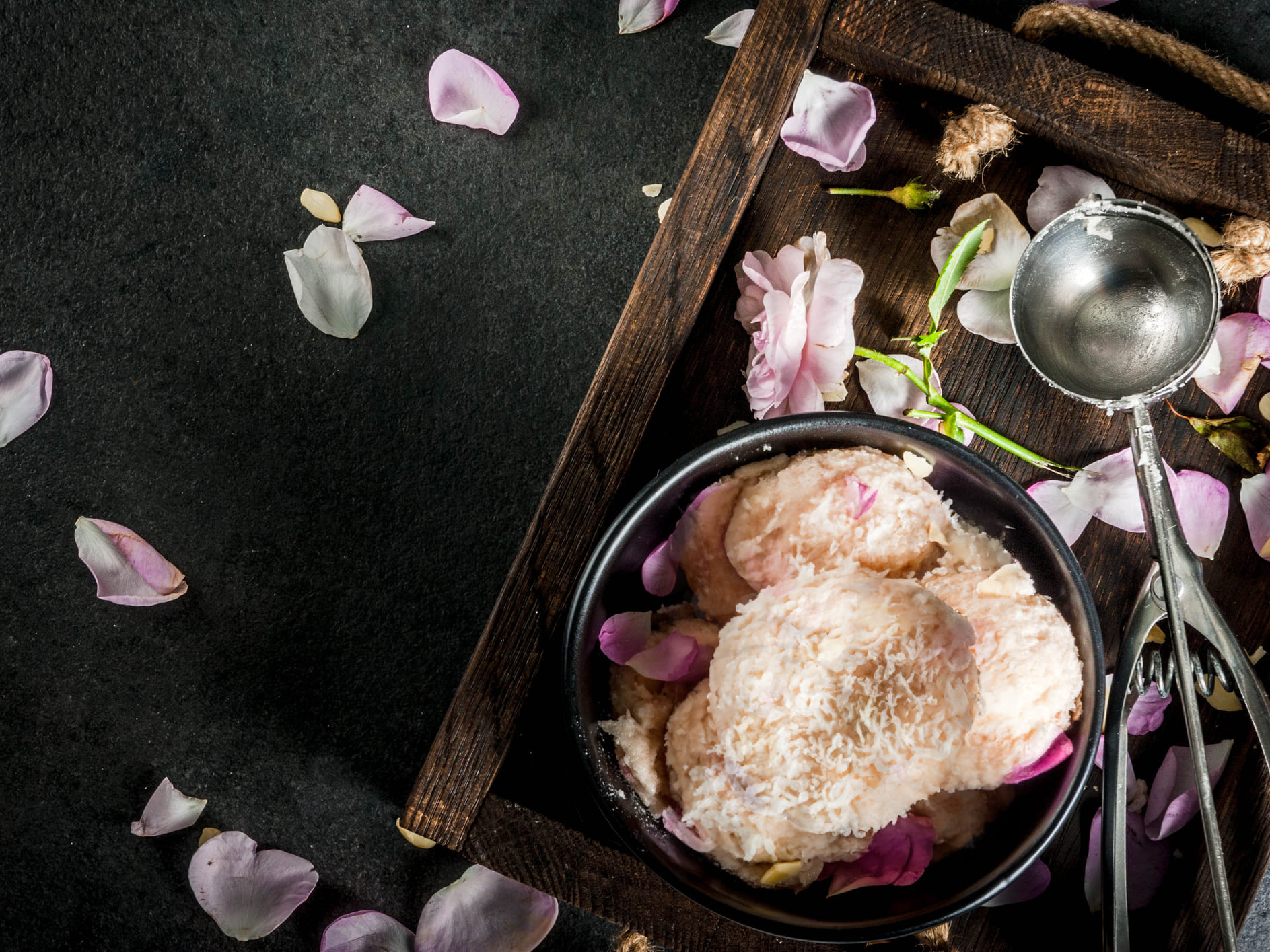 Kugeln rosafarbenes Eis mit Kokos in einer schwarzen Schale auf einem Holztablett mit Rosenblütenblättern