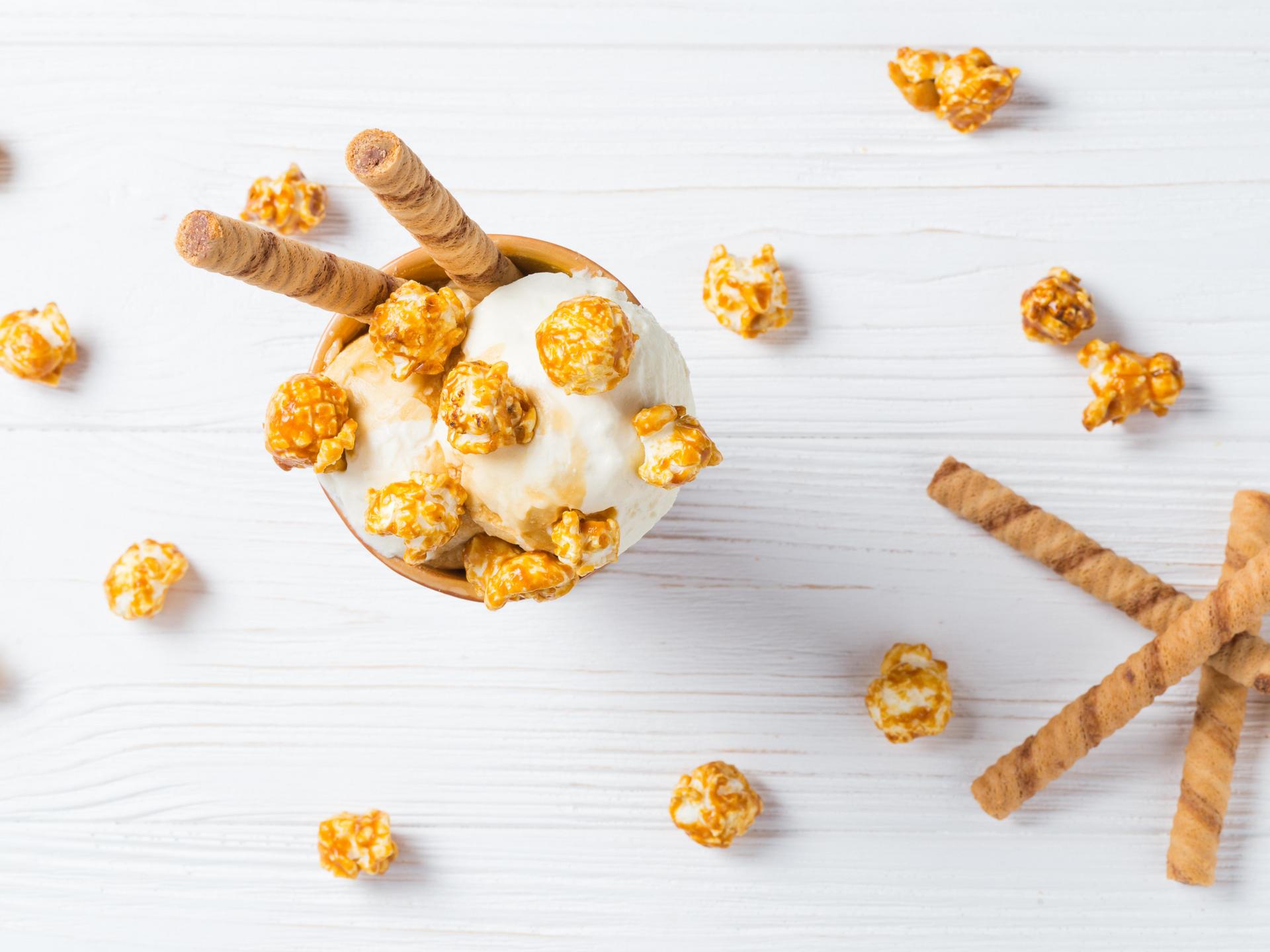 Schale Vanilleeis mit Karamell-Popcorn und Waffelröllchen auf einem weißen Holztisch