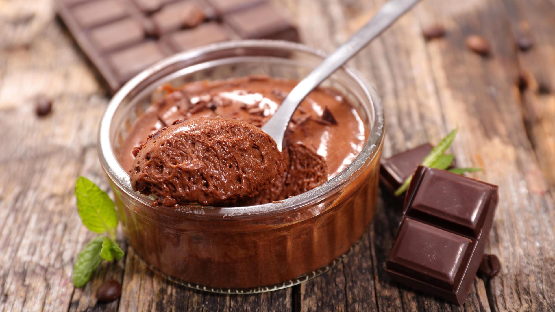 Bowl of chocolate mousse with a spoon, chocolate squares and mint leaves on a wooden table