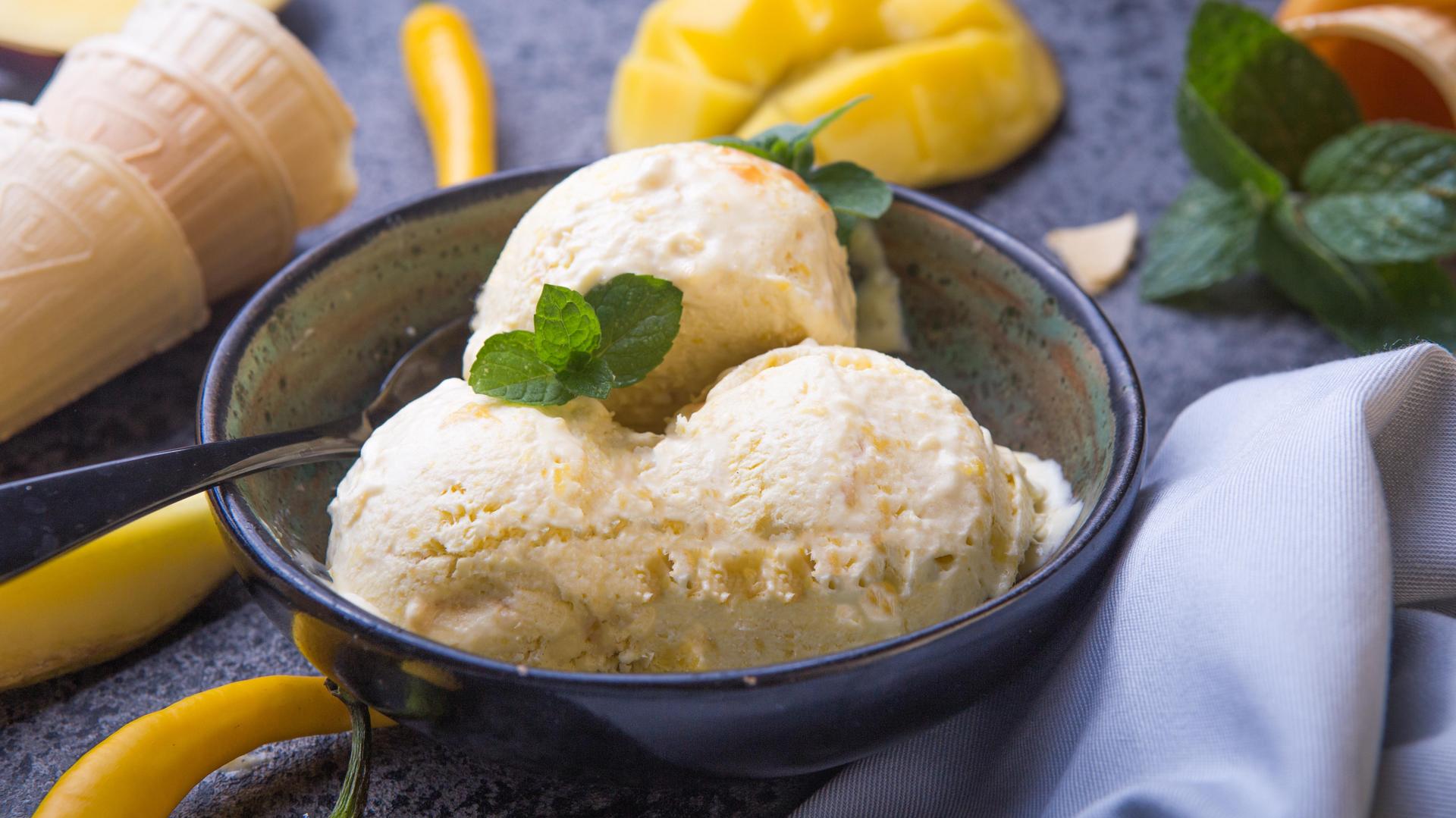 Bowl of mango ice cream scoops garnished with mint, surrounded by fresh mango pieces