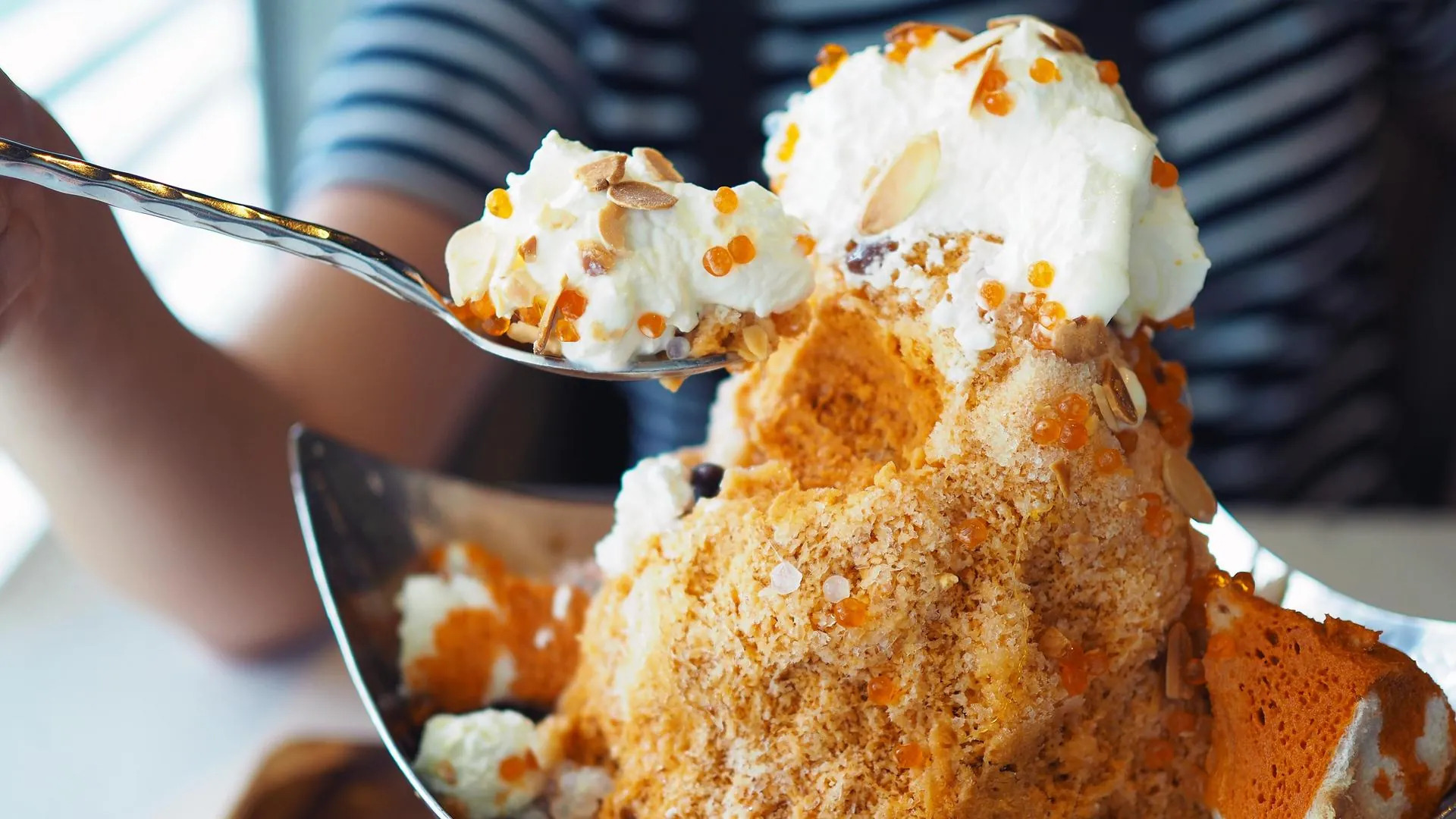 Spoon lifting a bite of shaved ice dessert topped with cream, popping boba and almond flakes