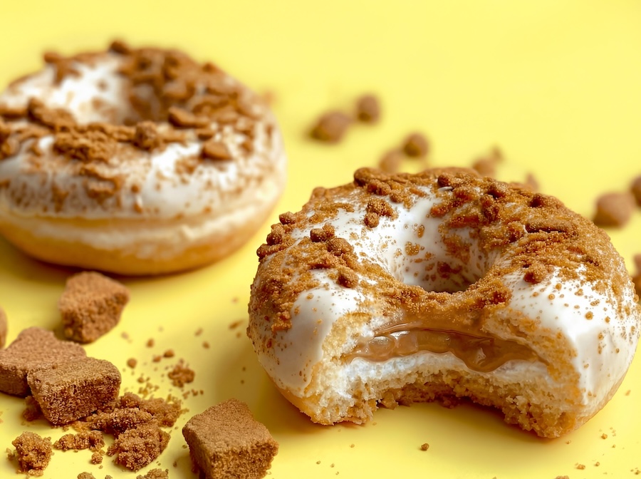 Zwei glasierte Donuts mit Keksbröseln, einer angebissen mit Karamellfüllung, vor gelbem Hintergrund