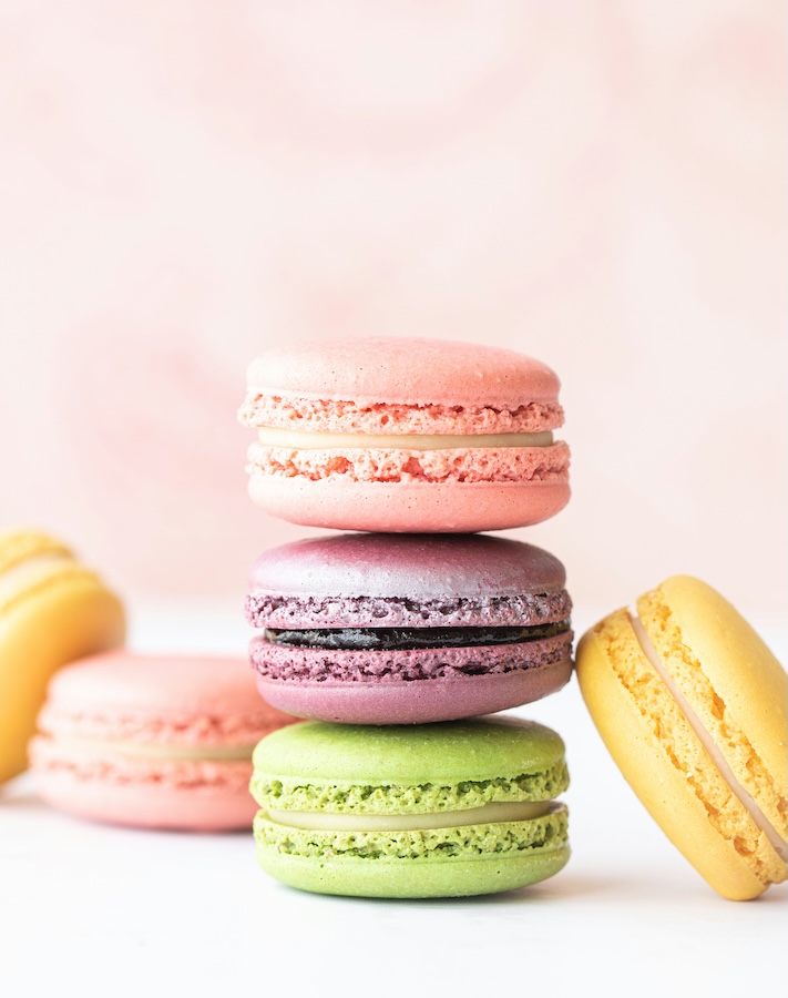 Stapel aus rosa, violetten und grünen Macarons, daneben gelbe Macarons vor rosa Hintergrund