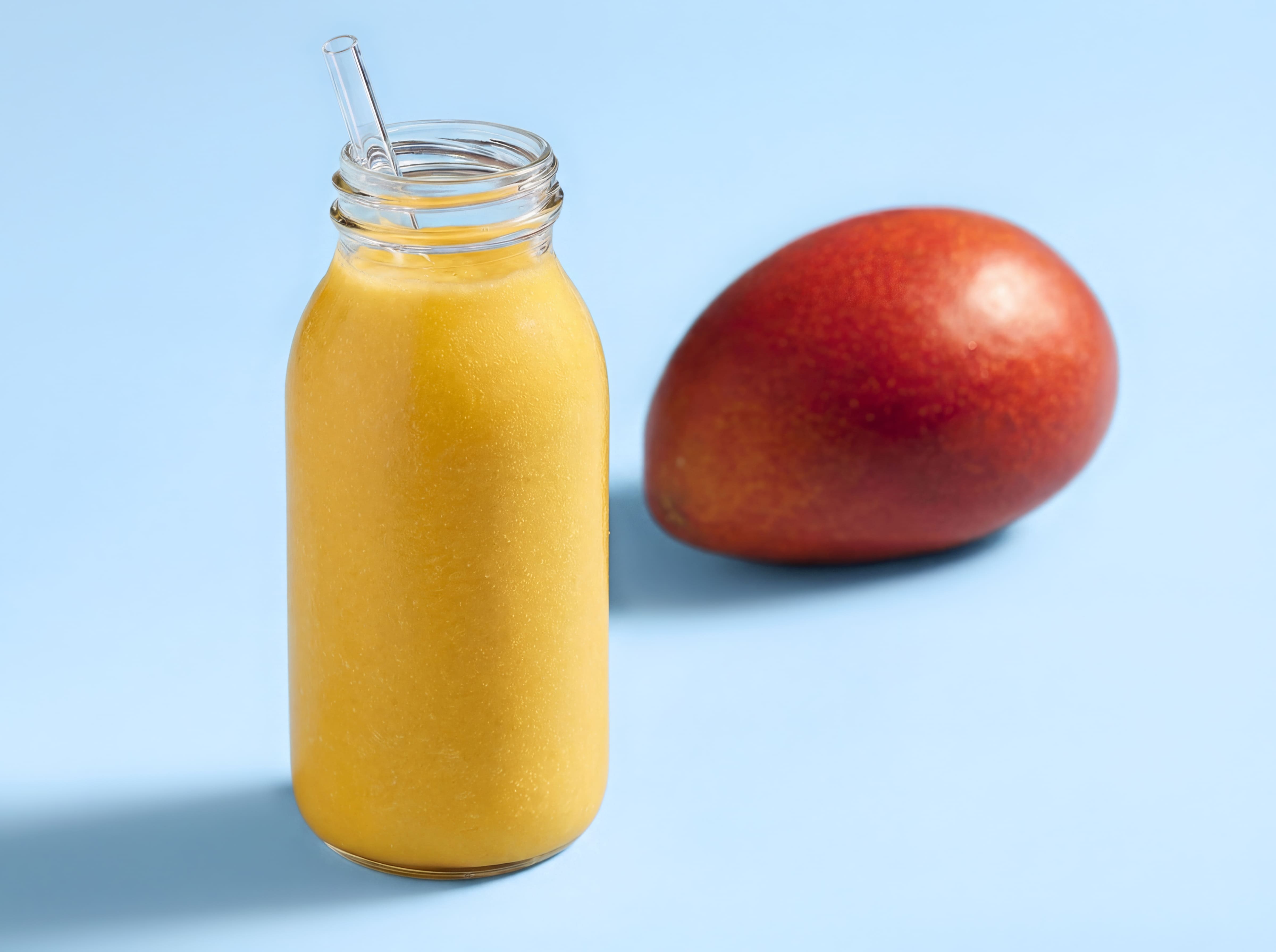 Glasflasche mit Mango-Smoothie und Strohhalm neben einer ganzen Mango vor hellblauem Hintergrund