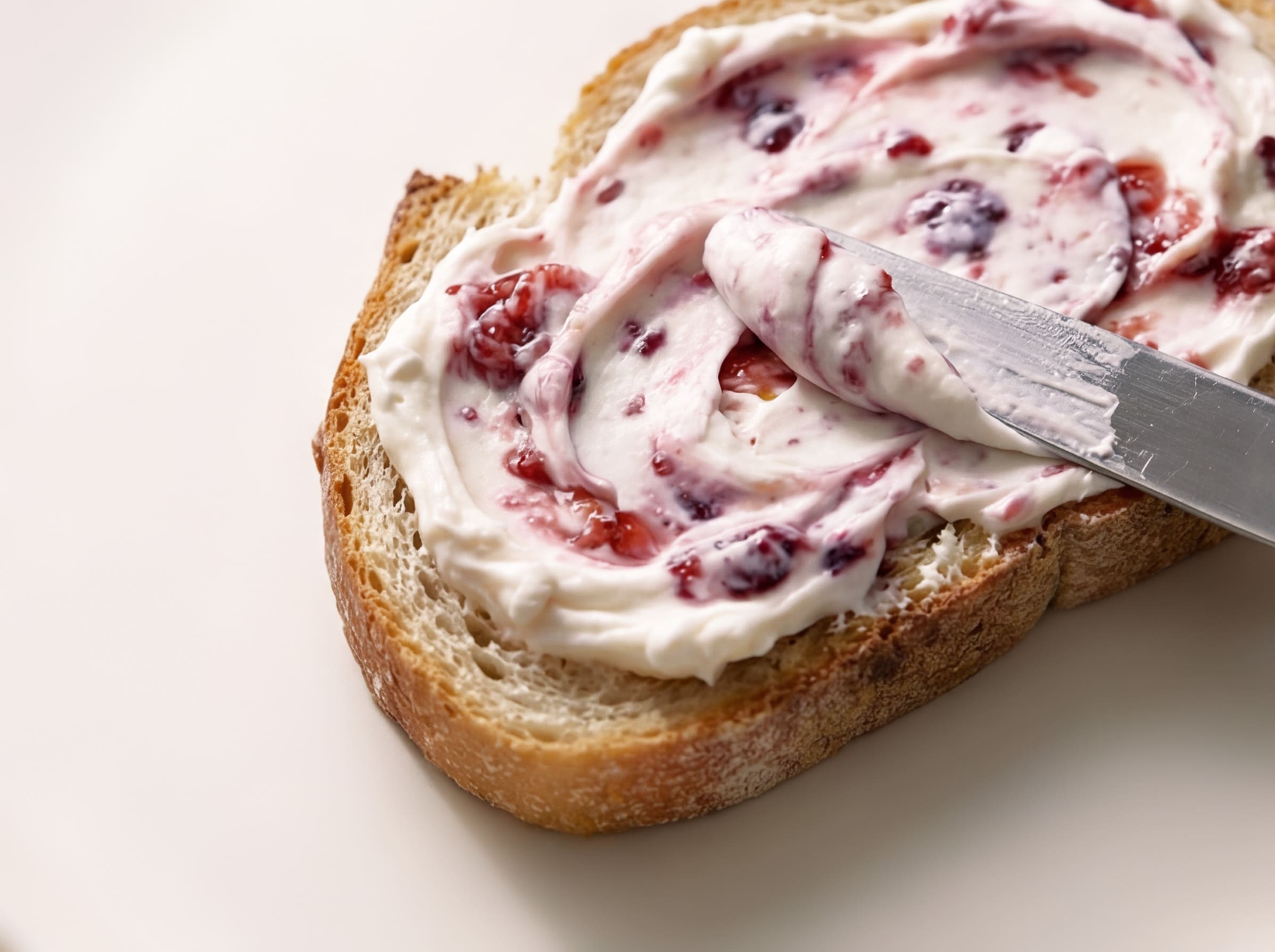 Messer streicht Frischkäse mit Beeren-Swirl auf eine Brotscheibe