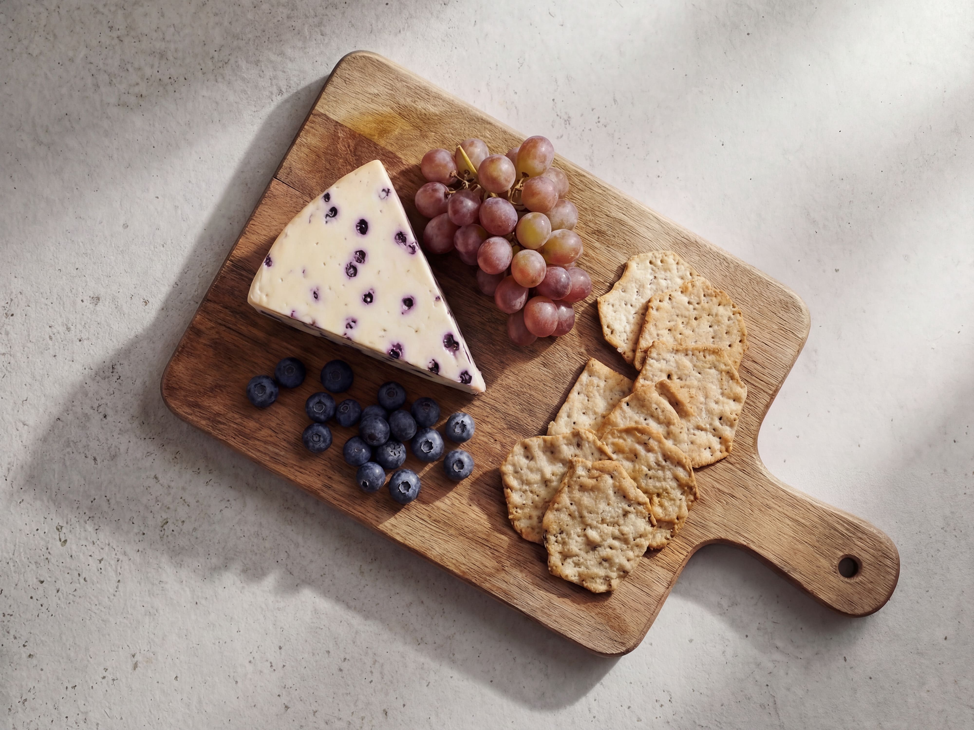 Stück Heidelbeerkäse auf einem Holzbrett mit frischen Heidelbeeren, roten Trauben und Crackern