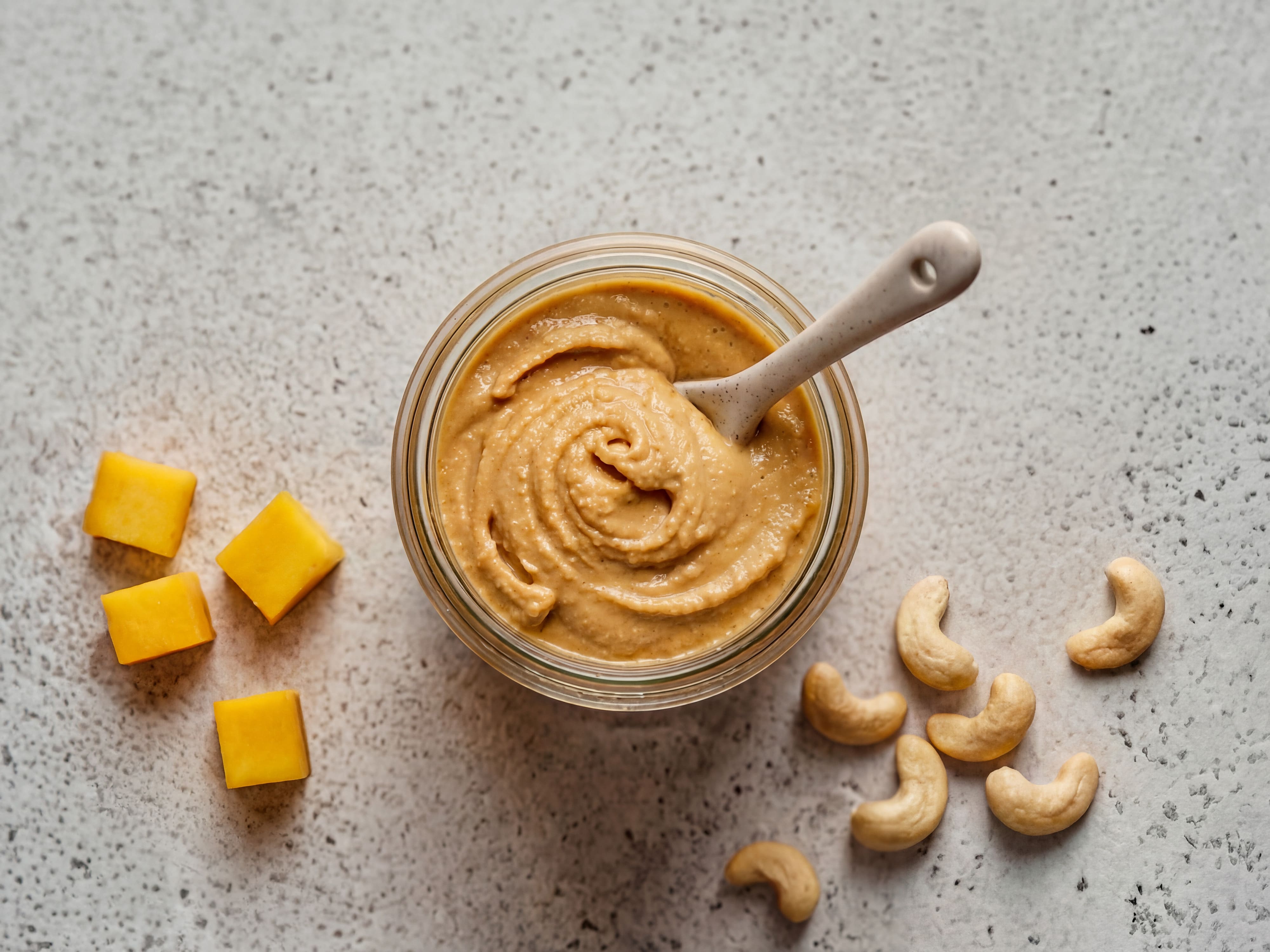 Glas mit cremigem Mango-Cashew-Aufstrich von oben, daneben Mangowürfel und Cashewkerne