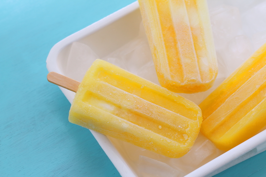 Gelbes Mango-Fruchteis am Stiel auf Eis in einer weißen Schale vor türkisem Hintergrund