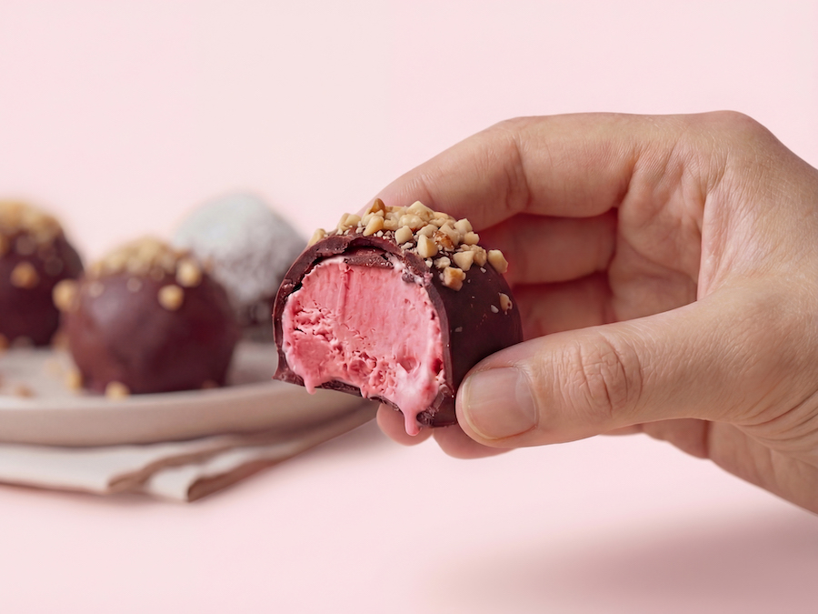 Hand hält ein angebissenes Eis-Praliné mit Schokoladenhülle, pinkem Fruchteiskern und gehackten Nüssen