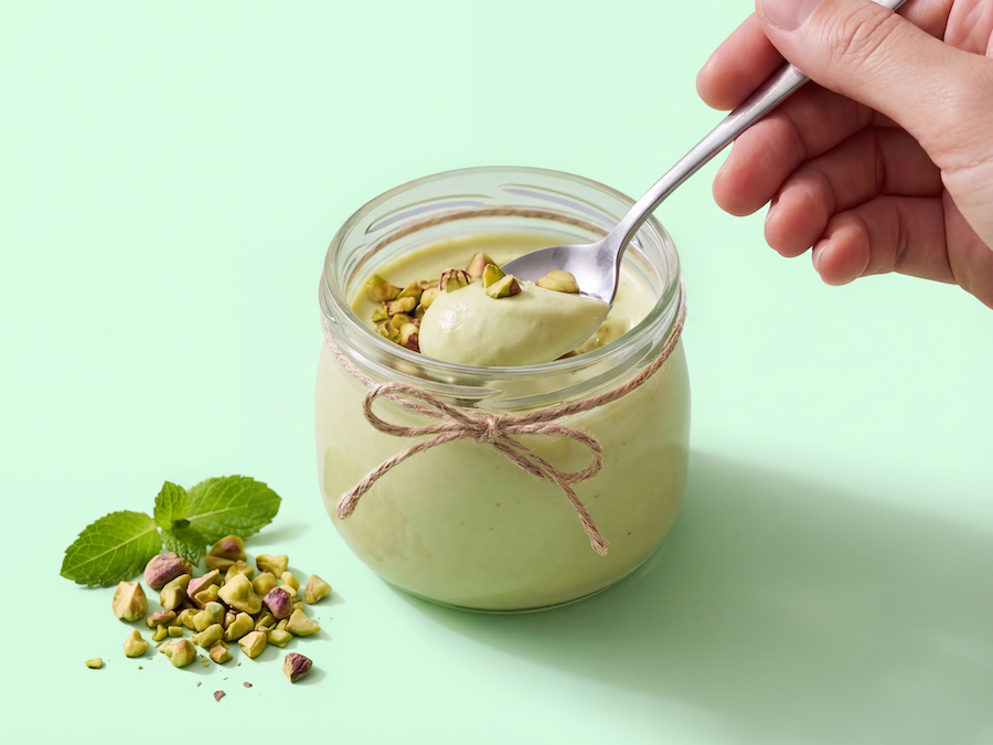 Hand löffelt Pistaziendessert aus einem kleinen Glas mit Schnurband, daneben gehackte Pistazien und Minze