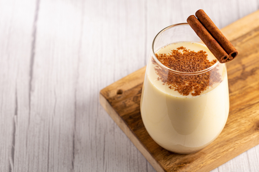 Glas Vanille-Milchshake mit Zimt bestäubt, zwei Zimtstangen am Rand, auf einem Holzbrett