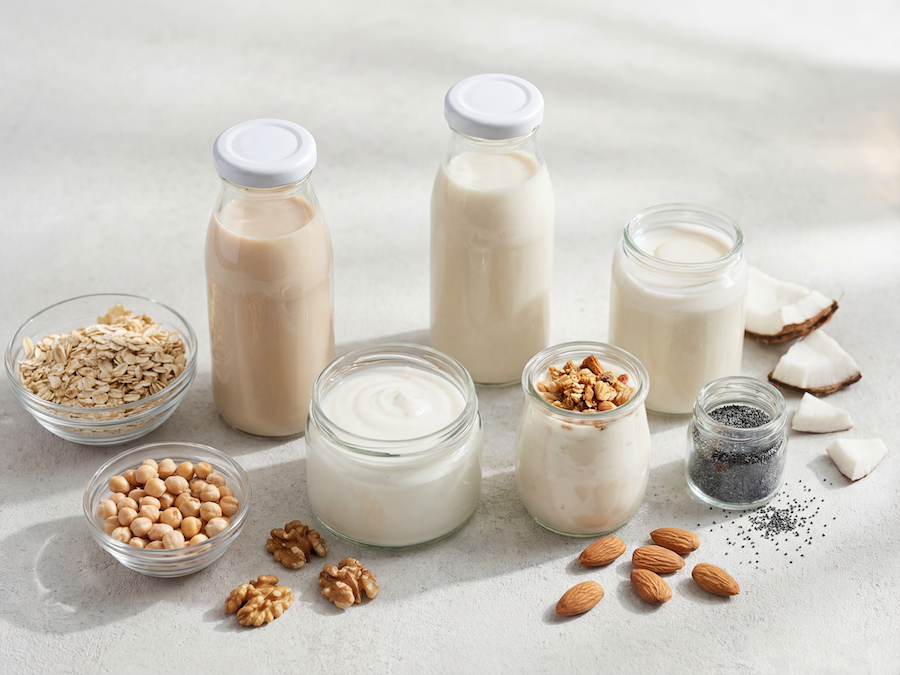 Flaschen und Gläser mit pflanzlichen Milch- und Joghurtalternativen, dazu Haferflocken, Kichererbsen, Nüsse und Kokos