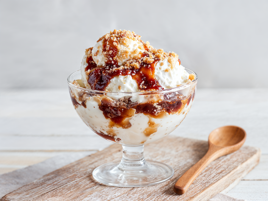 Glasschale mit Vanilleeis, Karamellsauce und knusprigen Keksstückchen, daneben ein Holzlöffel
