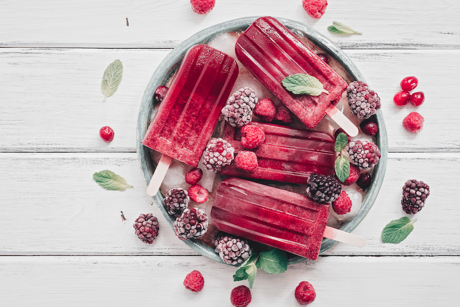 Rote Beeren-Eis am Stiel auf einem Tablett mit gefrorenen Brombeeren, Himbeeren, Cranberrys und Minze von oben
