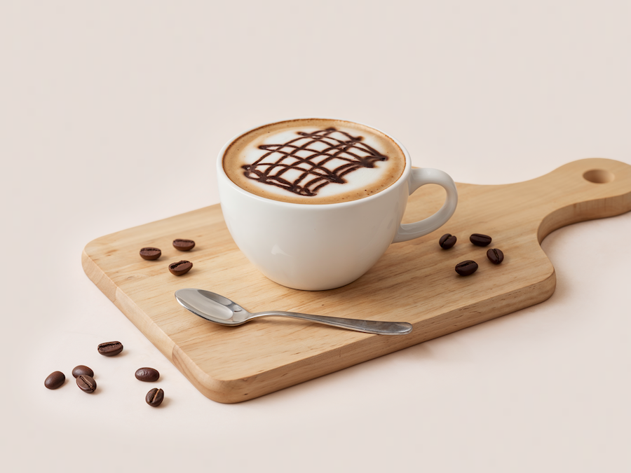 Cappuccino mit Schokoladensirup-Gitter auf dem Schaum, dazu Kaffeebohnen auf einem Holzbrett