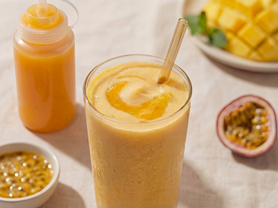 Mango-Maracuja-Milchshake im hohen Glas mit Strohhalm, daneben Fruchtsirup-Flasche, Maracuja und Mangowürfel