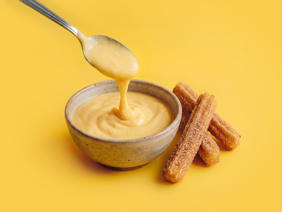 Löffel hebt cremigen Mango-Dip aus einer Schale, daneben Churros vor gelbem Hintergrund