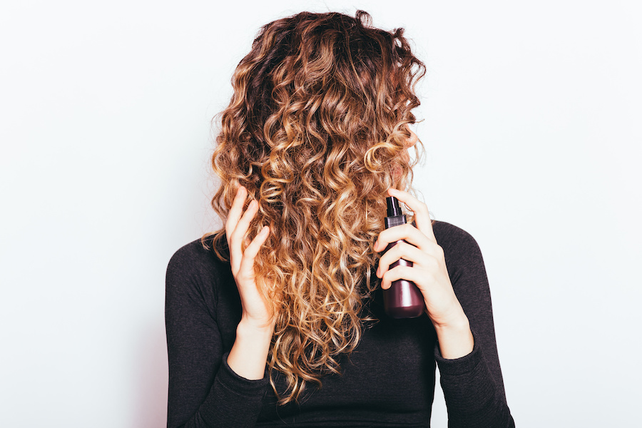 Frau mit langen Locken vor dem Gesicht hält eine Pumpsprayflasche mit Haarprodukt