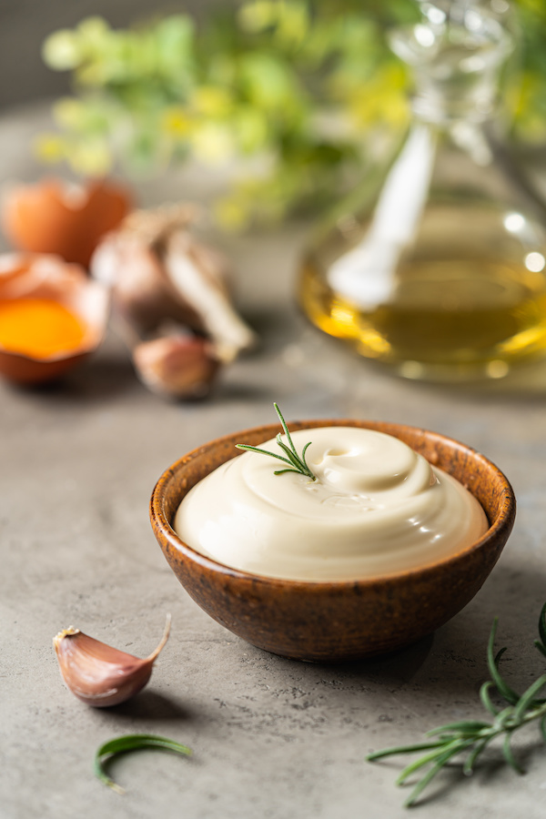 Holzschale mit cremiger Mayonnaise und Rosmarin, im Hintergrund Eier, Knoblauch und Öl