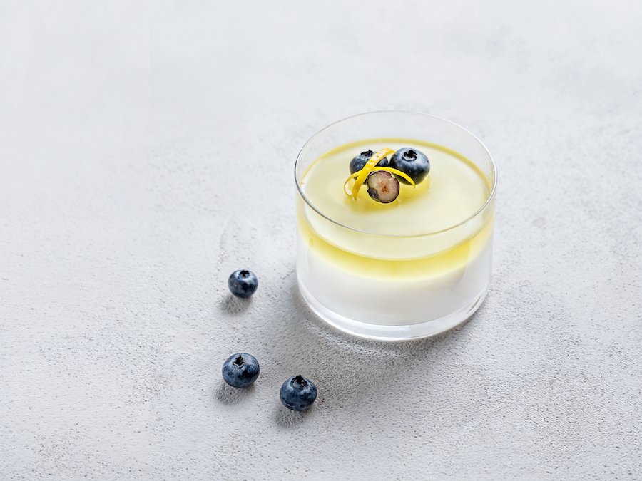 Glas Panna cotta mit Heidelbeeren und Zitronenzesten, daneben einzelne Heidelbeeren auf hellem Untergrund