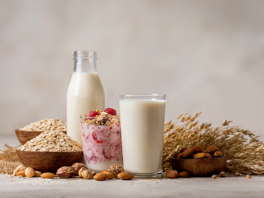 Flasche und Glas Haferdrink, Glas Beeren-Joghurtalternative mit Granola, umgeben von Haferflocken, Mandeln und Getreideähren