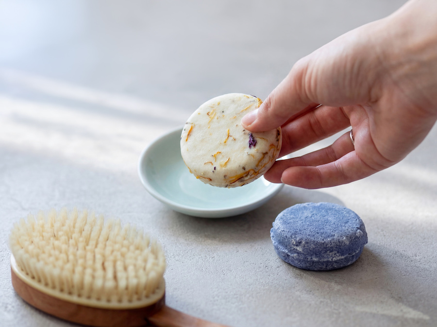 Hand nimmt ein festes Shampoo mit Blütenblättern, daneben ein lila Conditioner-Stück und Haarbürste