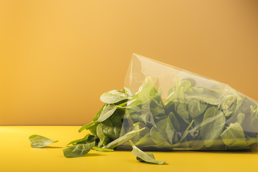 Frische Spinatblätter fallen aus einem durchsichtigen Obst- und Gemüsebeutel vor gelbem Hintergrund