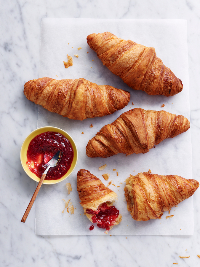 Goldbraune Croissants auf Backpapier, eines aufgebrochen, dazu ein Schälchen Erdbeermarmelade