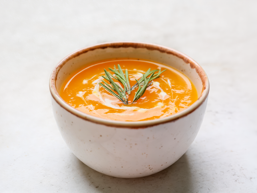 Schale mit oranger Cremesuppe und Rosmarinzweig auf hellem Hintergrund