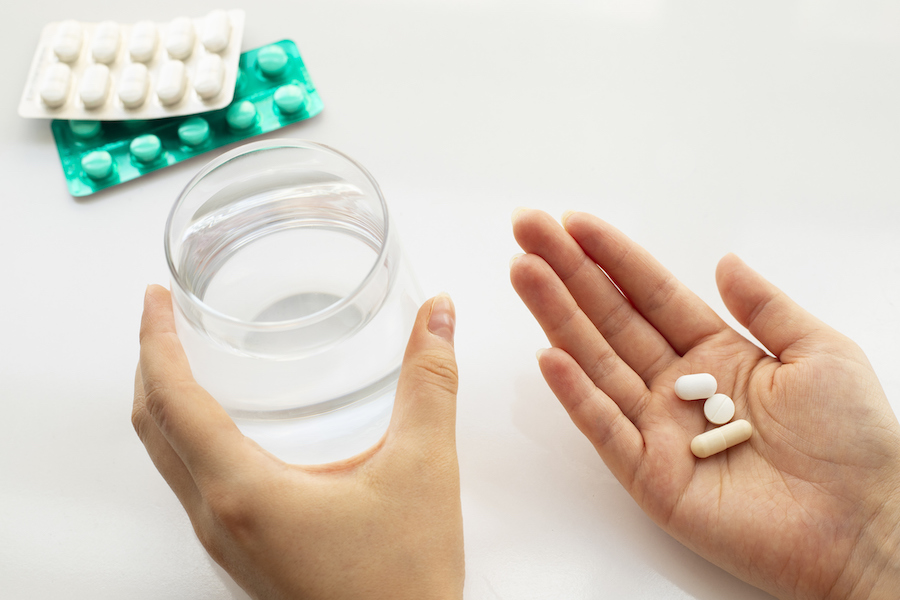 Hände halten ein Glas Wasser und drei Pillen, im Hintergrund Blisterpackungen