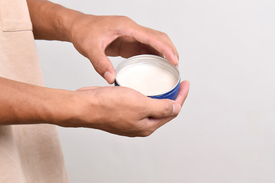 Hände halten eine offene Dose mit weißer Haarstyling-Paste