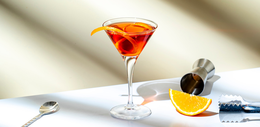 Orangeroter Likör-Cocktail im Martiniglas mit Orangenzeste, Orangenspalte und Barwerkzeug