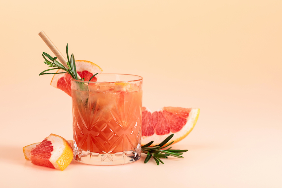 Glas mit Grapefruitdrink, Rosmarin und Grapefruitscheiben auf pfirsichfarbenem Hintergrund