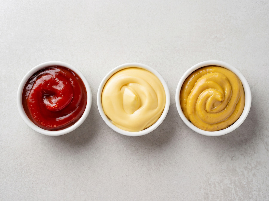 Drei Schälchen mit Ketchup, Mayonnaise und Senf von oben auf hellgrauer Fläche