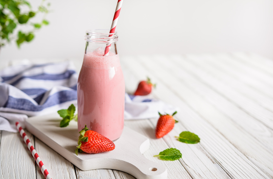 Flasche Erdbeer-Smoothie mit gestreiftem Strohhalm, frischen Erdbeeren und Minze auf einem weißen Brett