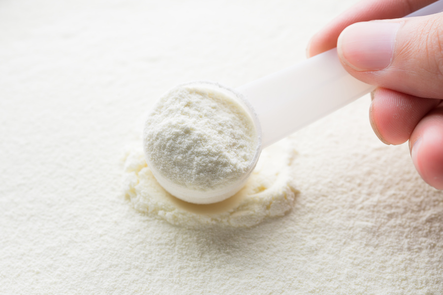 Hand hält einen Messlöffel Säuglingsmilchpulver über eine Fläche aus Milchpulver