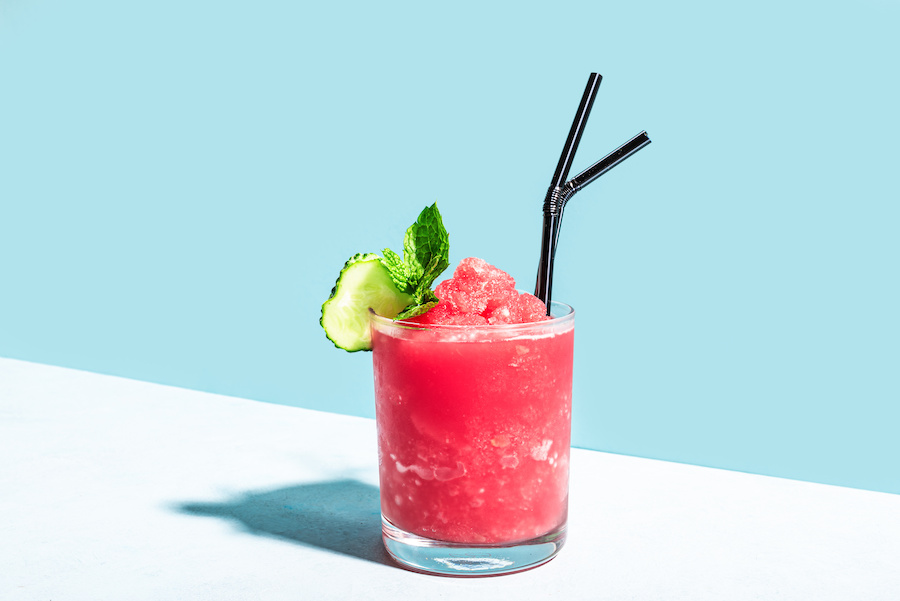 Glass of frozen watermelon cocktail with cucumber, mint and two black straws on a light blue background