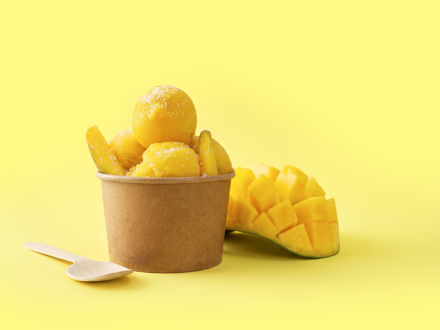 Scoops of mango sorbet in a paper cup with a wooden spoon and fresh mango on a yellow background