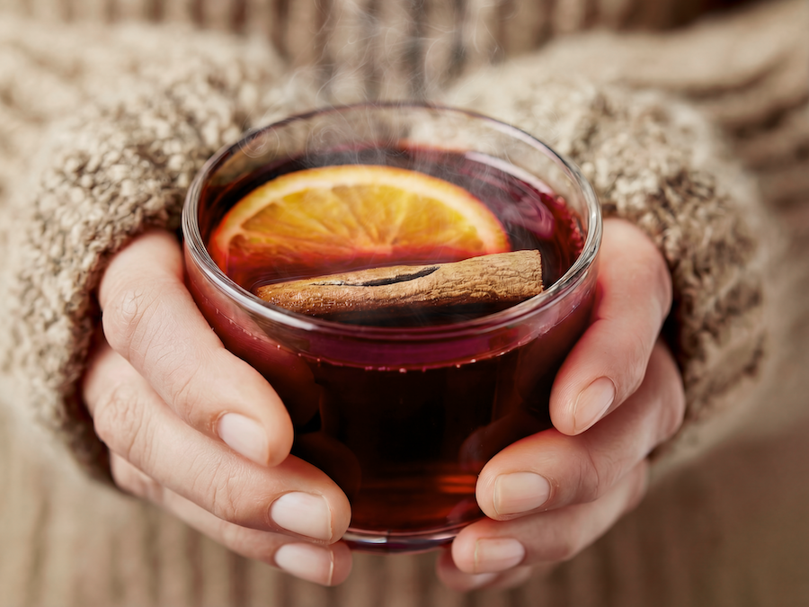 Hands in a knitted sweater cradling a glass of steaming mulled wine with an orange slice and cinnamon stick