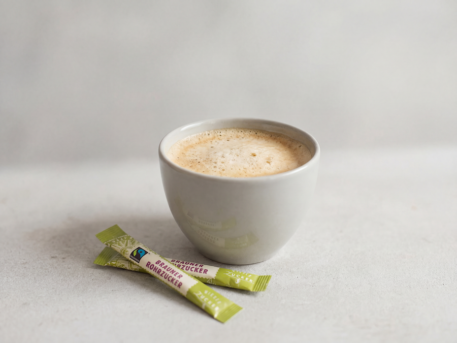 Tasse Kaffee mit Milchschaum und zwei Sticks Brauner Rohrzucker auf hellem Tisch